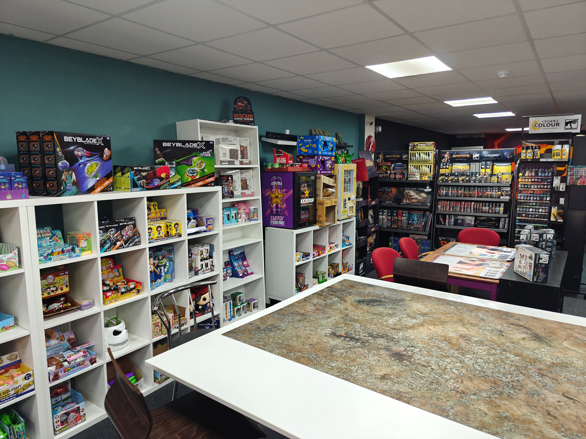A retail store interior with white shelving units filled with board games, toys, and collectibles, a large tabletop in the foreground, and shelving along the back wall.